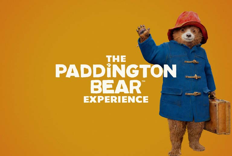 Paddington Bear wearing a blue coat and red hat, standing in a vibrant, colourful setting.